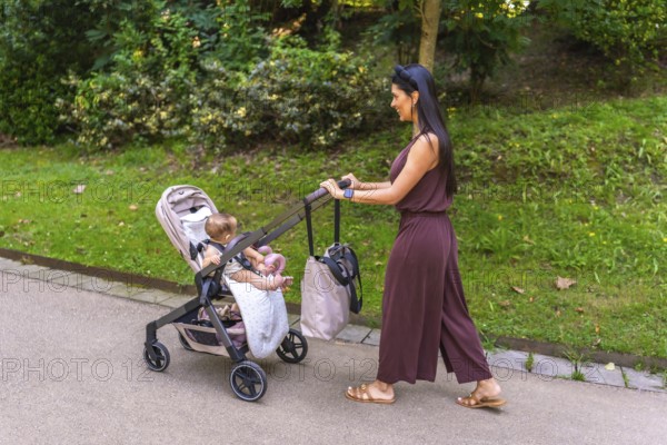 Young mother enjoying a sunny day in the park, happily pushing her baby girl in a stroller along a lush, green path