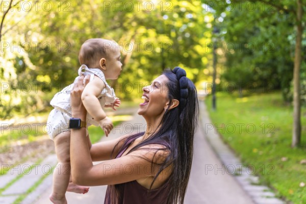 Smiling latin mother lifting her baby daughter while playing together in a sunny park, enjoying a beautiful day outdoors