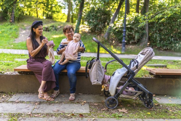 Grandmother holding baby girl while mother is eating and stroller is parked next to them on bench in park