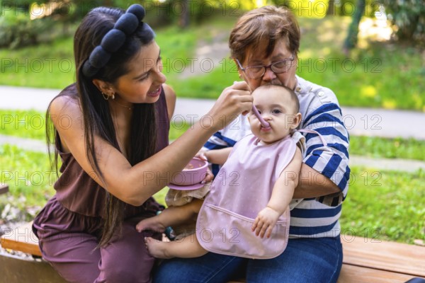 Grandmother holding baby girl while mother feeding her in the park, cherishing moments of family togetherness and joyful bonding