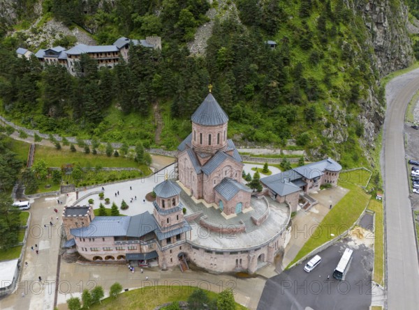 Large red brick church embedded in a wooded mountain landscape under a slightly cloudy sky, aerial view, Dariali Monastery complex, located in the Dariali Gorge of the Tergi River, Terek, Khevi Mountain Region, Georgian Military Road, Mtskheta-Mtianeti Province, High Caucasus, Georgia