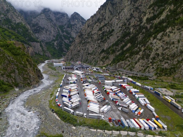 Car park full of lorries in a narrow mountain valley next to a river under a cloudy sky, aerial view, Larsi border crossing in Georgia towards Russia, Tergi river, Terek, Khevi mountain region, Georgian Military Road, Mtskheta-Mtianeti province, High Caucasus, Georgia