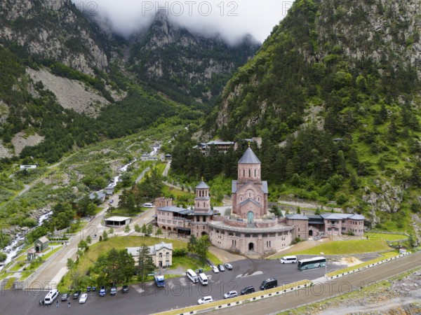 A large red brick church lies picturesquely in a mountainous valley under clouds, aerial view, Dariali Monastery complex, located in the Dariali Gorge of the Tergi River, Terek, Khevi Mountain Region, Georgian Military Road, Mtskheta-Mtianeti Province, High Caucasus, Georgia