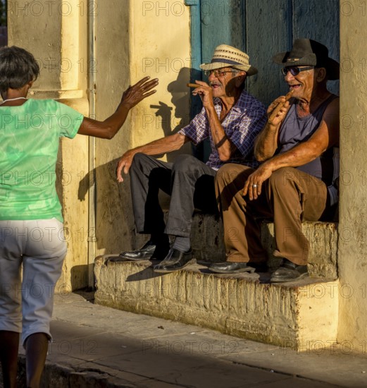 Two old Cubans smoke cigars and sit on a staircase in the warm light of the sunset, they greet a passer-by in a green T-shirt, Trinidad, Cuba, Sancti Spíritus, Sancti Spíritus