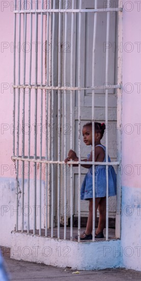 Little coloured girl behind window bars in a pink house, street of Trinidad, Cuba, Sancti Spíritus