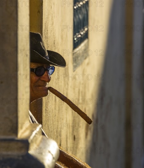 An old Cuban smokes a cigar and sits on a staircase in the warm light of the sunset, Trinidad, Cuba, Sancti Spíritus, Sancti Spíritus
