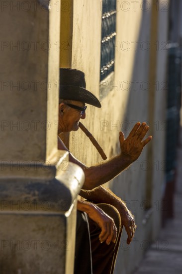 An old Cuban smokes a cigar and sits on a staircase in the warm light of the sunset, Trinidad, Cuba, Sancti Spíritus, Sancti Spíritus