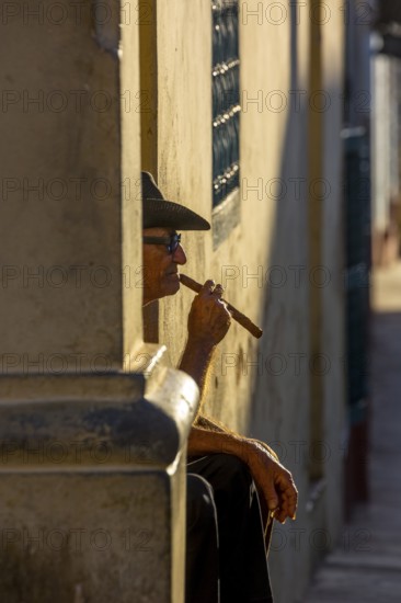 An old Cuban smokes a cigar and sits on a staircase in the warm light of the sunset, Trinidad, Cuba, Sancti Spíritus, Sancti Spíritus