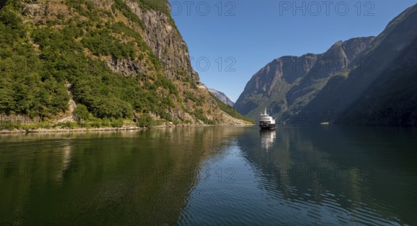 An approaching excursion boat on a beautiful fjord reflecting the light, surrounded by rocks partly overgrown with trees, above a bright blue sky. Gudvangen, Sogn og Fjordane, Norway, Scandinavia, Europe, Gudvangen, NOR, travel, tourism, destination, sightseeing, sea voyage, Hurtigruten, Hurtigruten trip, tourism, Norwegian, Scandinavian, tourist, cruise, adventure trip, mountains, tourist trip, boat trip, river, rocks, tourist ship, water, waters