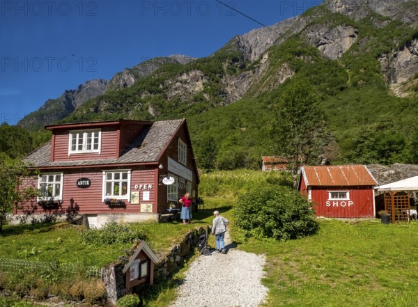 Tourists at mountain hut station, red wooden houses, gravel path, mountains, meadow, blue sky, Gudvangen, Sogn og Fjordane, Norway, Scandinavia, Europe, NOR, travel, tourism, destination, sightseeing, sea voyage, Hurtigruten, Hurtigruten trip, tourism, Norwegian, Scandinavian, tourist, cruise, adventure trip, houses, stones, rocks, trees