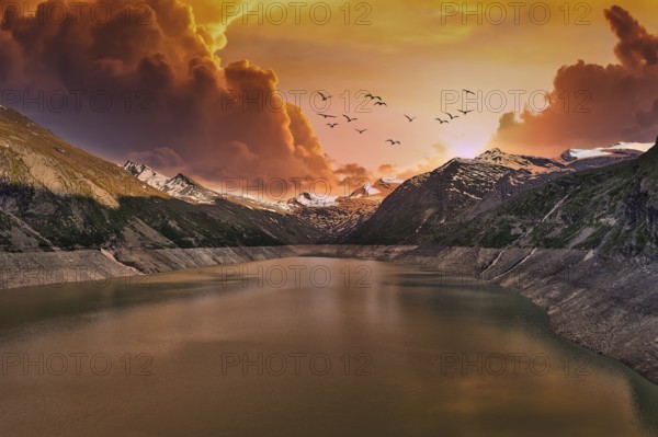Dramatic sunset over a mountain lake with flying birds and clouds, Mattmark Reservoir, Saas Almagell, Saas Grund, Canton Valais, Switzerland