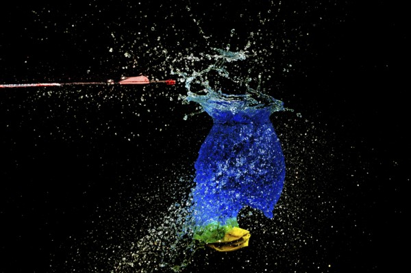 A blue water balloon bursts bubbling, with an arrow and yellow elements in a haunting scene, high speed shot, water balloon, arrow
