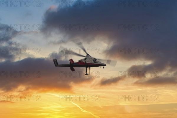 A helicopter flies in front of a dramatic sky with clouds and sunset, transport helicopter, timber transport, Saas Grund, Canton Valais, Switzerland