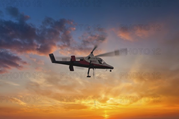A helicopter hovers elegantly in front of a brightly coloured sunset sky, transport helicopter, timber transport, Saas Grund, Canton Valais, Switzerland