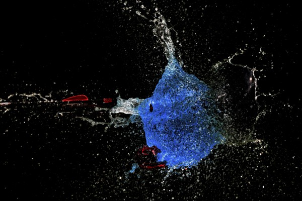 A blue water balloon bursts with force while an arrow shoots through it, surrounded by a dark background, high-speed shot, water balloon, arrow