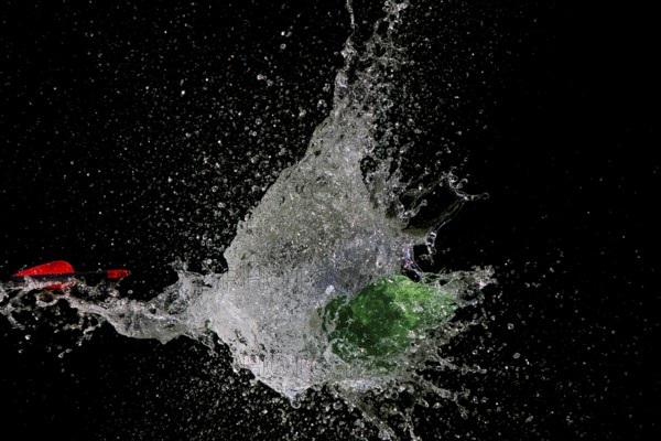 A green water balloon bursts after arrow penetration, causing water to splash, in front of a dark background, high-speed shot, water balloon, arrow