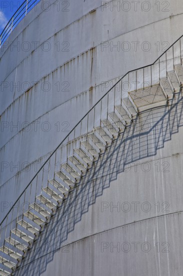 A long metal ladder stretches along a large cylinder building with shadows in the background, elevated tank, chemical, company, Grenzach- Wyhlen, Baden-Württemberg, Germany