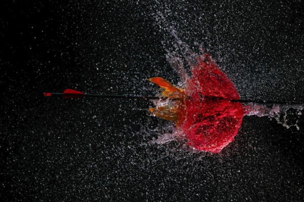 An arrow pierces a red water balloon and creates an impressive water explosion against a dark background, high-speed shot, water balloon, arrow