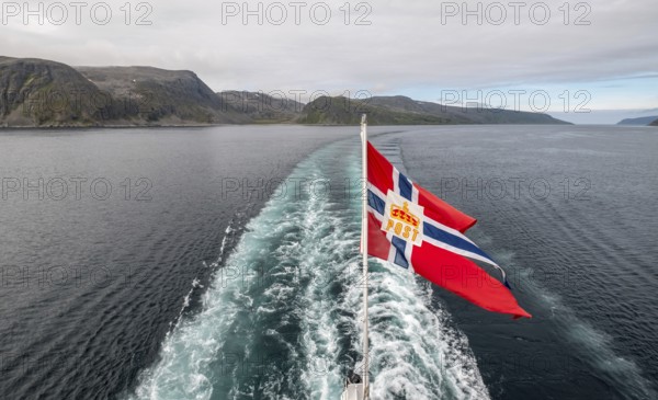The post symbol on a Norwegian flag of a Hurtigruten ship, foaming spray, Honningsvåg, Finnmark, Norway, Scandinavia, Europe, adventure travel, fjord, flags, tourism, Honningsvåg, Hurtigruten, Hurtigruten trip, cruise, NOR, mail ship, travel, destination, sea trip, sightseeing, tourism, Norwegian, Norwegian flag, Scandinavian, touristic