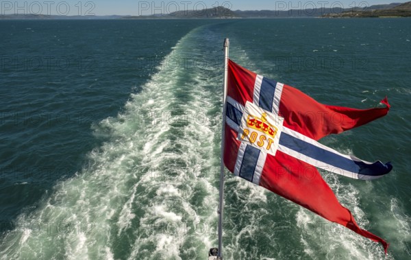 The post symbol on Norwegian flag of a Hurtigruten ship, foaming spray, Trondheimfjord, Agdenes, Trøndelag, Norway, Scandinavia, Europe, adventure trip, fjord, flags, tourism, Hurtigruten, Hurtigruten trip, cruise, NOR, mail ship, travel, destination, sea trip, sightseeing, tourism, Trøndelag, Norwegian, Scandinavian, touristic