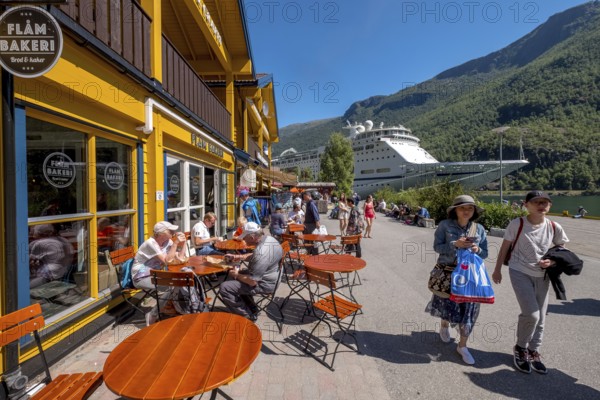 Tourists in Flåm, FABRIK KUTSALG FACTORY OUTLET, café, yellow wooden house, excursion ship, fjord, mountain, trees, blue sky, Sogn og Fjordane, Norway, Scandinavia, Europe, Flåm, NOR, travel, tourism, destination, sightseeing, sea voyage, Hurtigruten, Hurtigruten voyage, tourism, Norwegian, Scandinavian, tourist, cruise, adventure trip, pedestrian, river, forests, shipping, shops, shopping, shopping, signs, flags, tables, chairs, people, windows, shops, balconies
