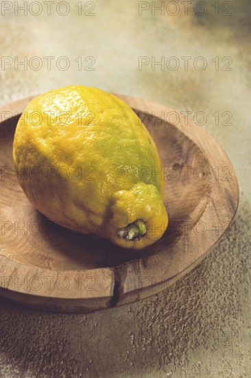 A vibrant lemon sits on a rustic wooden plate, showcasing its uneven texture and vivid color. The background features soft natural textures, creating a warm kitchen atmosphere