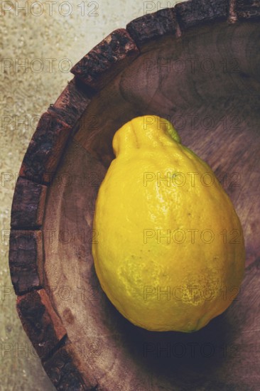 A vibrant yellow lemon sits alone in a wooden bowl with a rough rim, highlighting its natural texture against a muted background, evoking freshness and simplicity