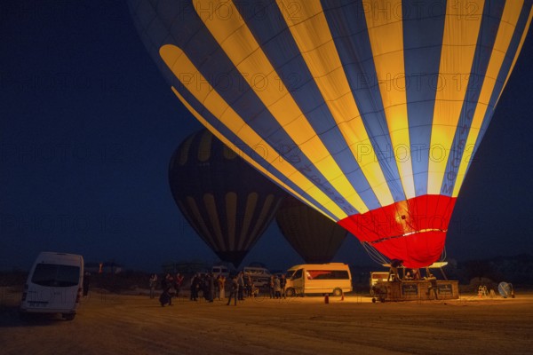 Hot air balloons at night with people and vehicles, bright colours in contrast to the dark sky, Göreme National Park, Göreme Tarihî Millî Parki, Nevsehir province, Nevsehir, Cappadocia, Cappadocia, Kapadokya, Cappadocia, Central Anatolia, Turkey