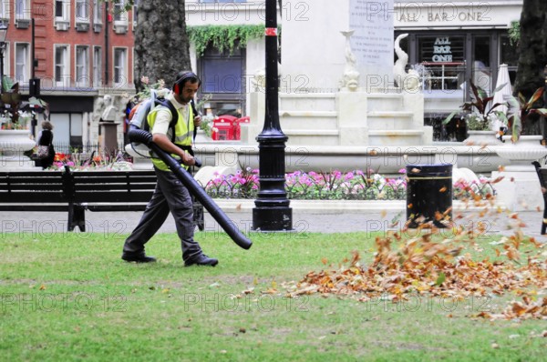 A worker uses a leaf blower to remove leaves in a municipal park, London, England, United Kingdom