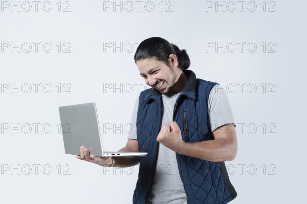 Male celebrating with fist in the air holding a laptop on white background. Young man celebrating holding a computer on isolated background