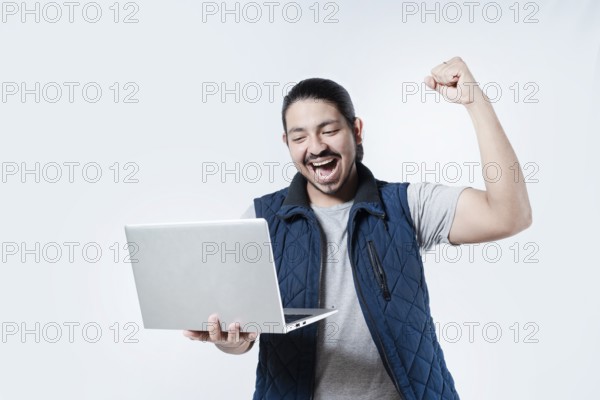 Young man celebrating holding a computer on isolated background. Male celebrating with fist in the air holding a computer on white background
