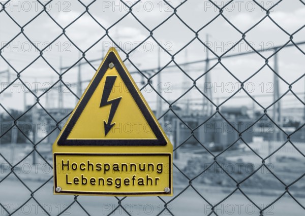 Yellow german high voltage warning sign on a mesh wire fence in front of a blue toned electrical substation in Southern Germany