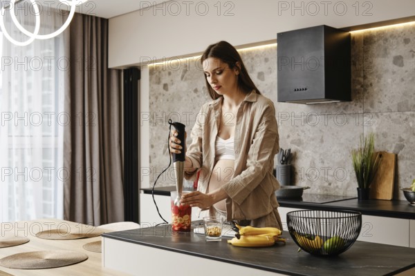 In a stylish kitchen, a pregnant woman blends fresh fruits like bananas and strawberries. Sunlight fills the space as she focuses on creating a nutritious smoothie, promoting a healthy lifestyle
