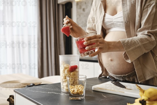 A pregnant woman is creating a nutritious smoothie in a stylish kitchen. She layers strawberries and bananas in blender container while standing by a cutting board with pine nuts