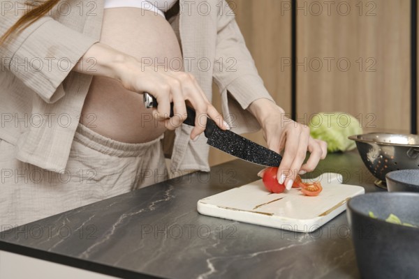 A woman in advanced pregnancy cuts fresh tomatoes on a chopping board in a stylish kitchen. She wears a comfortable outfit and focuses on preparing a nutritious salad while surrounded by ingredients