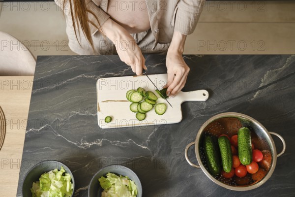A pregnant woman with long hair skillfully slices cucumbers on a cutting board in a stylish kitchen. Fresh vegetables are organized nearby, creating a vibrant and inviting atmosphere for meal prep