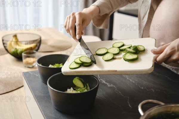 A person slices cucumbers on a wooden cutting board, adding them to two black bowls filled with salad ingredients