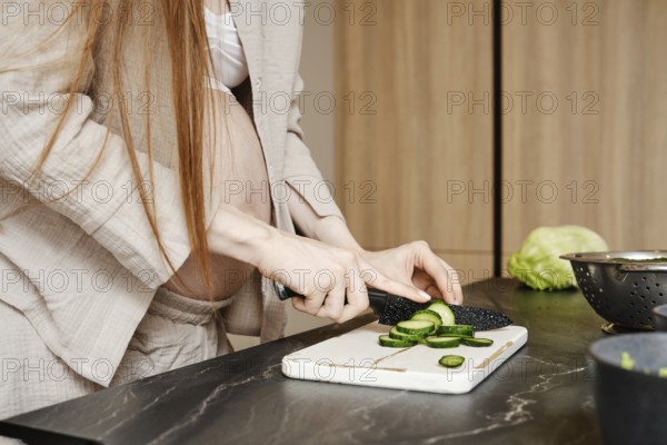 In a stylish kitchen, a pregnant woman with long hair cuts cucumbers on a white cutting board. She wears casual clothes and shows focus as she prepares healthy food, surrounded by fresh produce