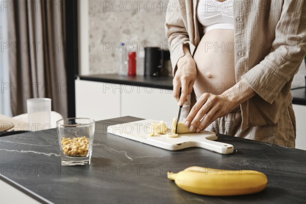 A pregnant woman slices a banana while standing at a countertop. A glass of nuts and a banana are visible, emphasizing healthy eating during pregnancy