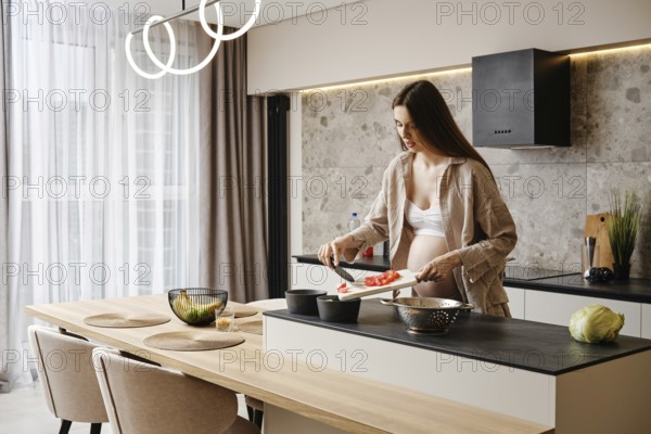 A pregnant woman with long hair is preparing a meal in a contemporary kitchen. She is cutting vegetables, showcasing healthy cooking. Natural light fills the room, enhancing a calm atmosphere