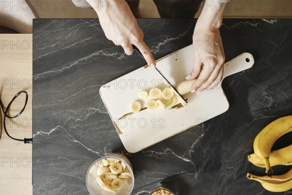 A person slices fresh bananas on a white cutting board in a well-lit kitchen. The countertop features a bowl of bananas and a glass jar. Sunlight streams in, creating a warm atmosphere