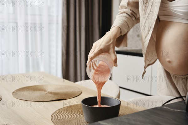 A pregnant woman pours a vibrant pink smoothie from a glass jar into a black bowl in a contemporary kitchen. Natural light fills the room, highlighting her focus on nutrition