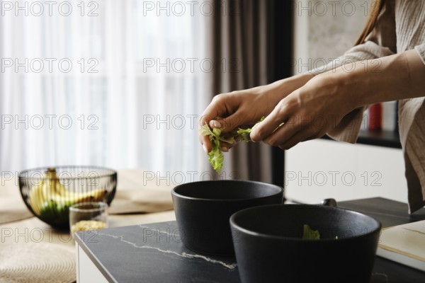 Hands gently tear lettuce leaves while preparing a fresh salad. The kitchen features contemporary design elements with soft lighting and a stylish arrangement of ingredients in the background