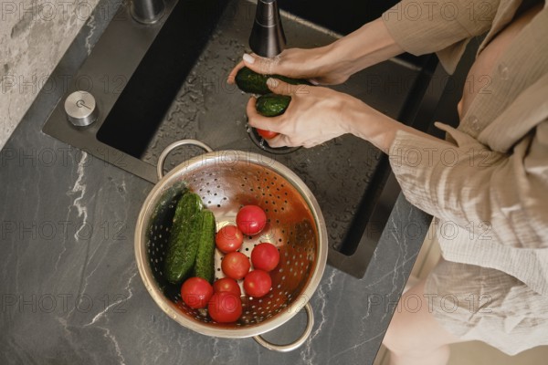Hands scrub cucumbers under running water at a sleek kitchen sink. A colander nearby holds ripe tomatoes and cucumbers, showcasing a vibrant assortment of fresh produce