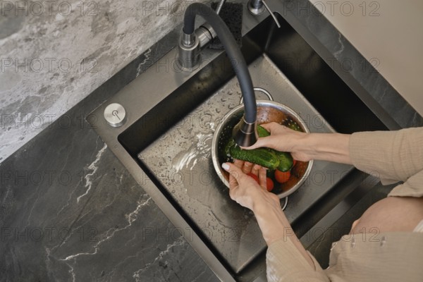 In a contemporary kitchen, a person is rinsing fresh vegetables under a stylish faucet. The dark countertop contrasts with the vibrant colors of cucumbers and tomatoes in a bowl