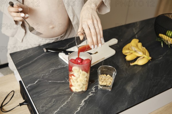 A person pours strawberry syrup over a mix of fruits, including bananas and strawberry, in a glass. The kitchen is bright, showcasing healthy ingredients on a dark marble countertop