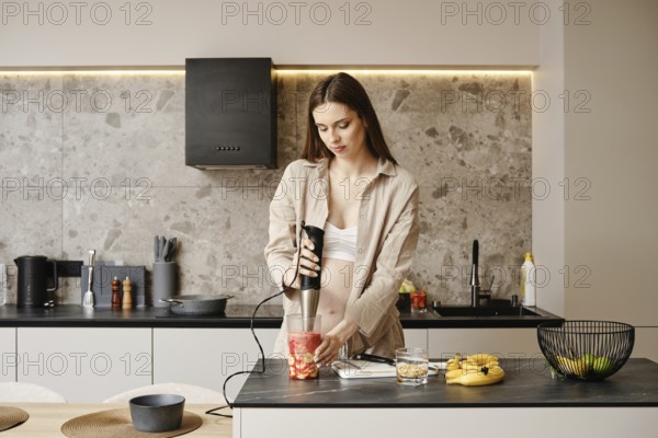 A pregnant woman stands in a contemporary kitchen using a handheld blender to prepare a fruit smoothie. Fresh ingredients are scattered across the counter, creating a vibrant, healthy atmosphere