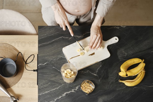A person with a visible baby bump is cutting bananas on a wooden cutting board in a modern kitchen. Fresh bananas and chopped pieces are arranged neatly, highlighting the preparation for a snack