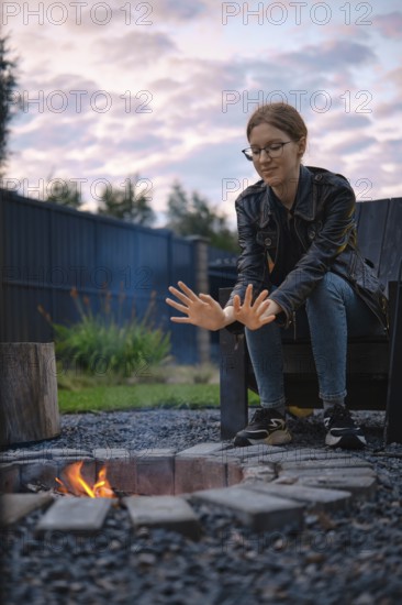 A young woman sits on an outdoor chair near a fire pit, warming her hands in the glow of the flames as dusk settles over a suburban backyard. Surrounding greenery adds a serene touch