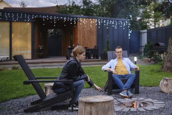 Father and his daughter sit by a fire pit in a backyard, engaging in conversation as the sun sets. Young woman stirs the coals in a campfire